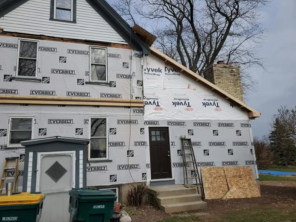 House exterior under construction, covered in Tyvek, with a wooden roof frame.