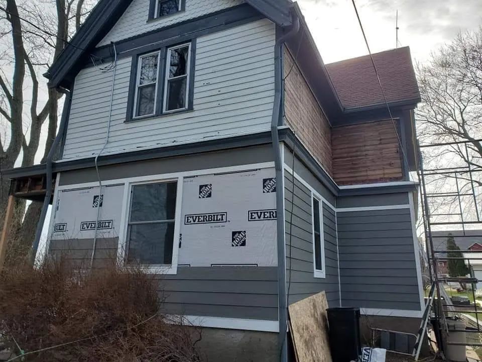 House exterior during renovation. Gray siding, partially covered with building wrap. Red roof.