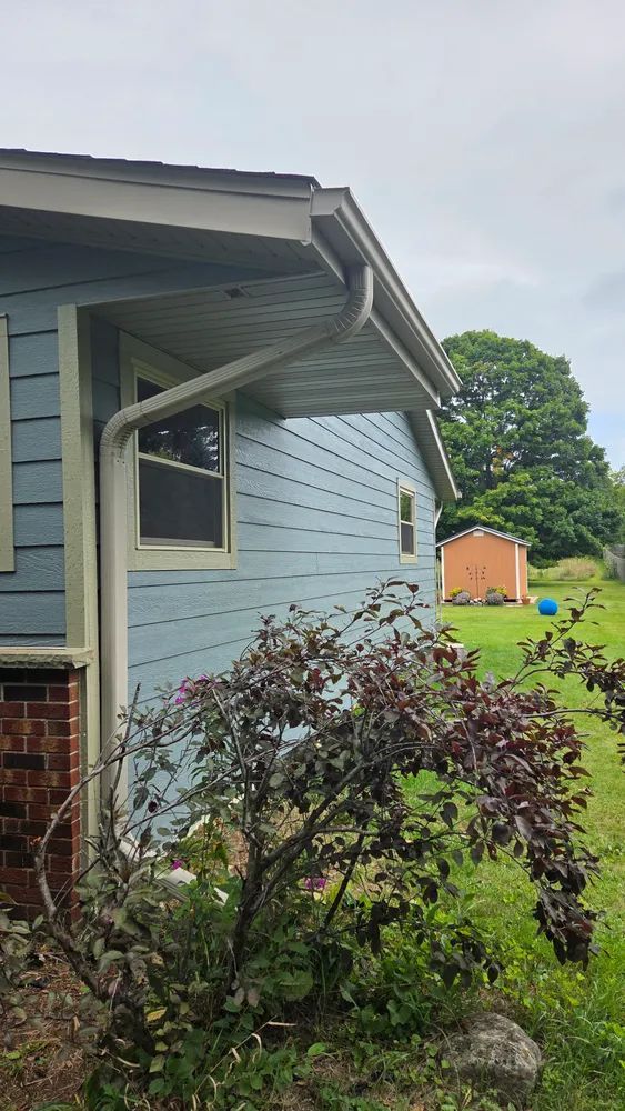 Light blue house with white trim, gutters, and a small shed in the background. Bushes in foreground.
