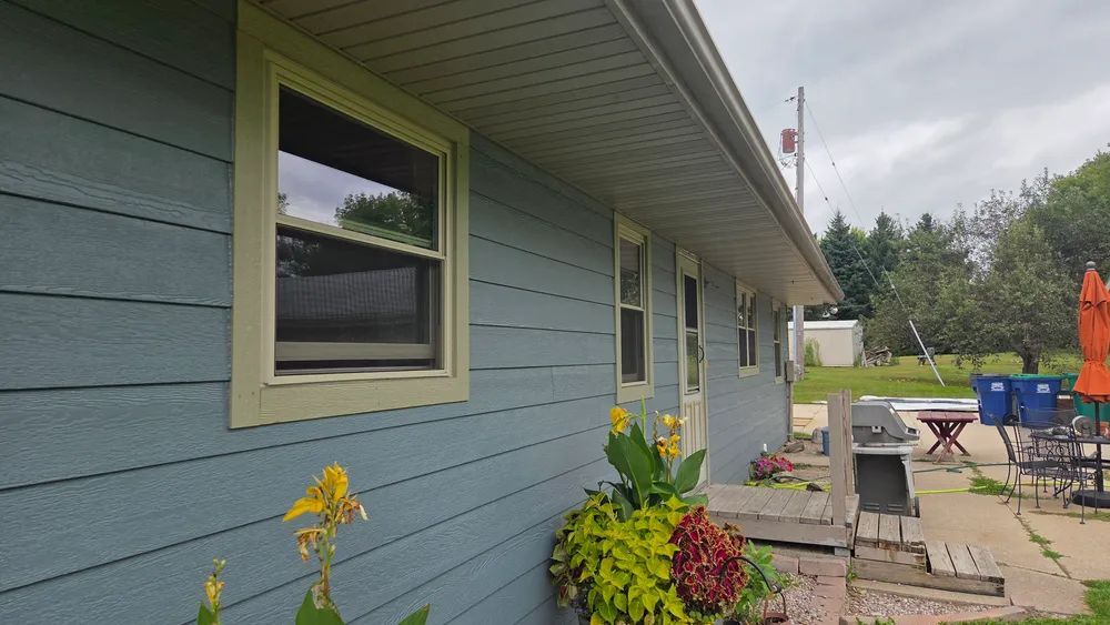 Blue-sided house with green trim and windows, a flower planter, and a patio with furniture and an umbrella.