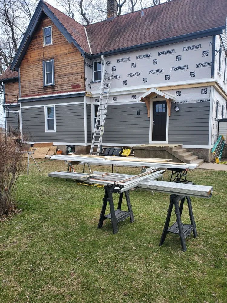 House exterior under renovation; gray siding, old wood siding, ladder, lumber on sawhorses, green grass.