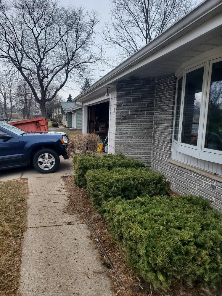 Blue car parked next to a house with a garage and green bushes. Sidewalk in front. Overcast day.