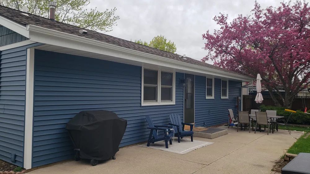 Blue house exterior with patio, grill, chairs, and blooming pink tree.