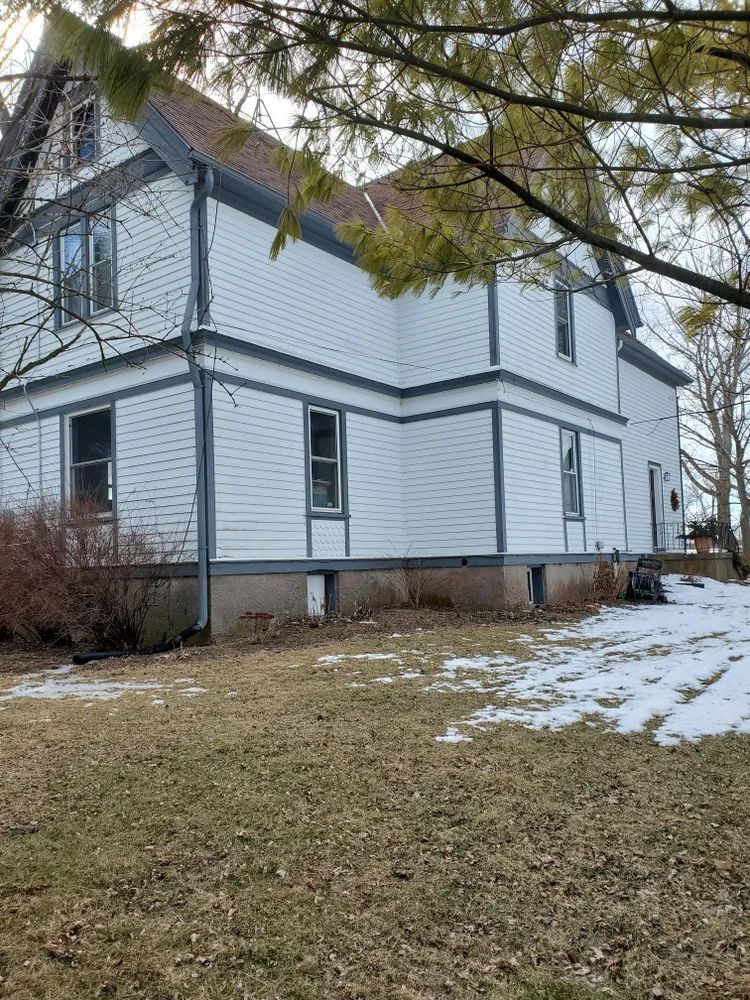 Two-story white house with blue trim, built on a brown foundation. Brown grass and patches of snow in the yard.