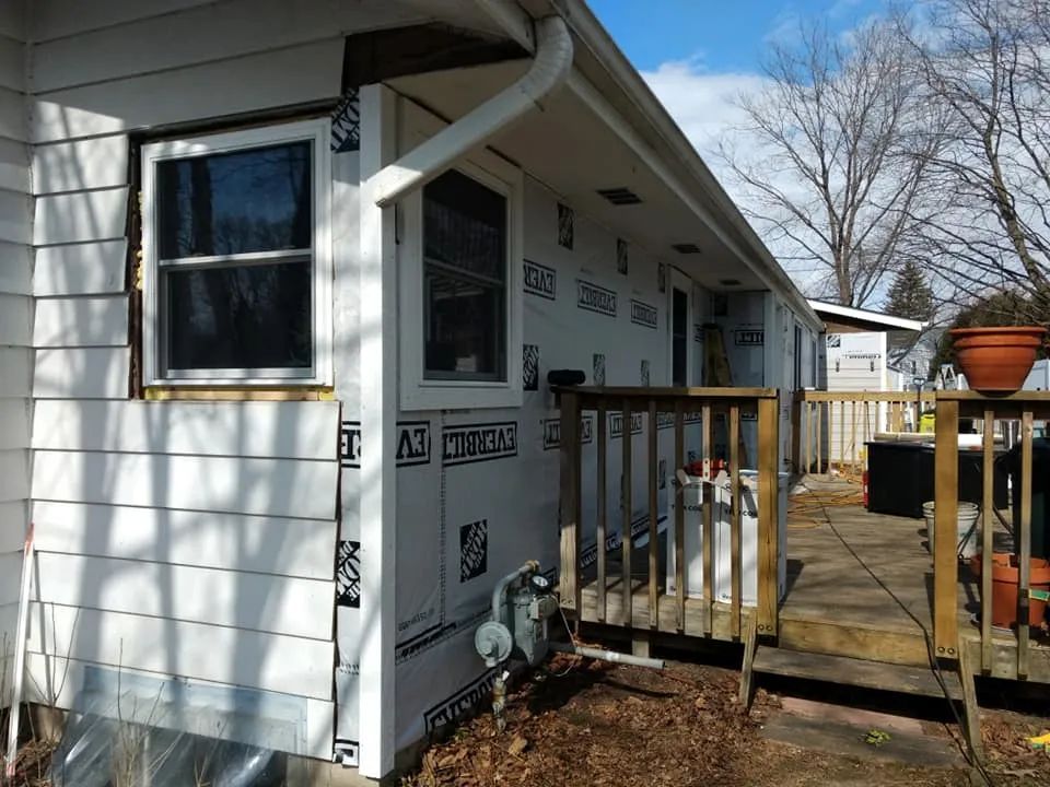 House exterior partially covered in white siding with black wrap, next to wooden deck.
