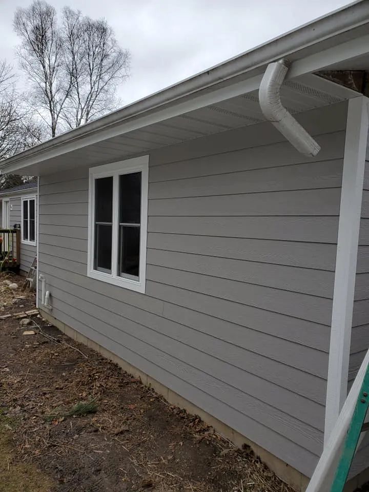 Gray siding on a house with white window frames and gutters, a cloudy sky, and a yard with dead leaves.