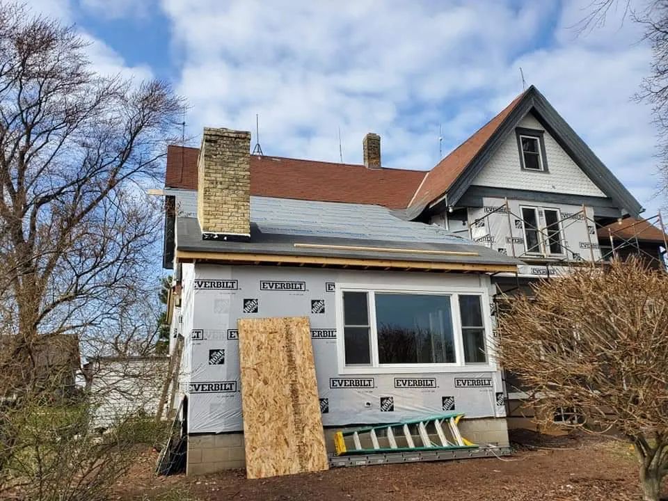 House under construction with tarpaper roof, new siding, window, chimney, ladder, and OSB board.
