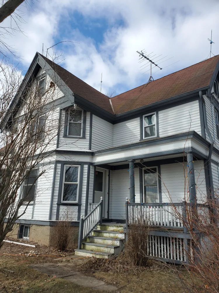 Two-story white house with gray trim, a porch, and a brown roof against a cloudy blue sky.