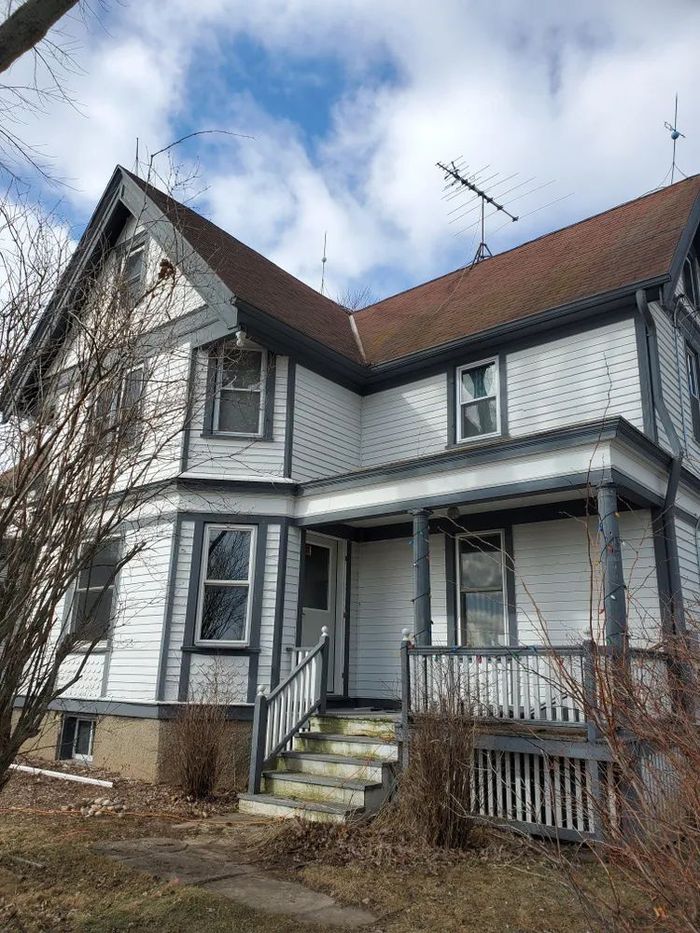 Two-story white house with gray trim, a porch, and a brown roof against a cloudy sky.