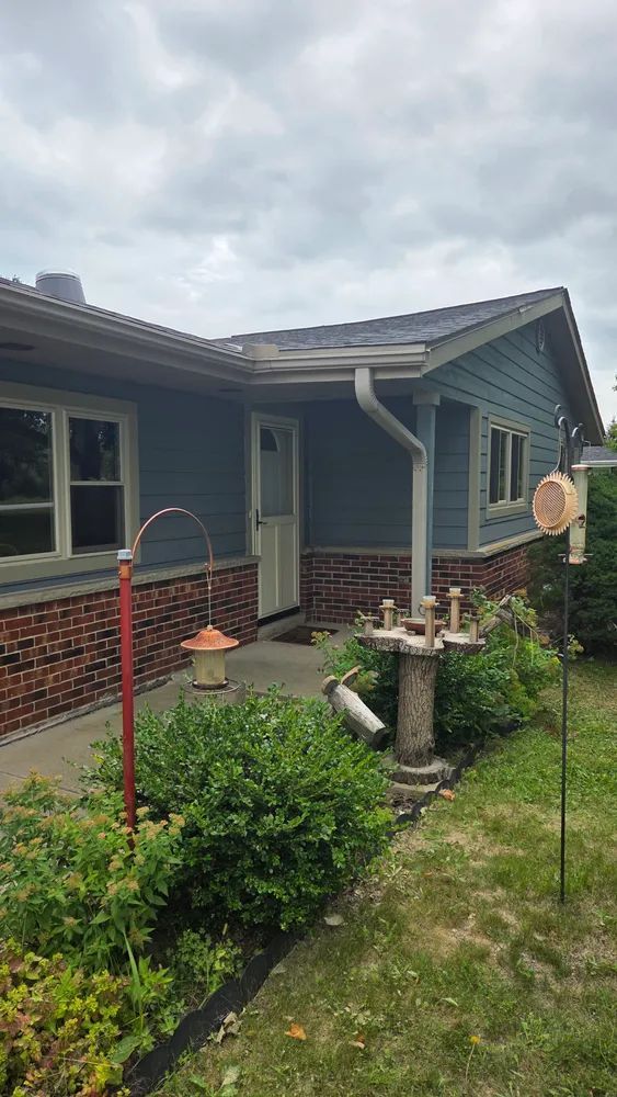 A house with blue siding, brick base, and a white door, with bird feeders and lush green bushes in the front yard.