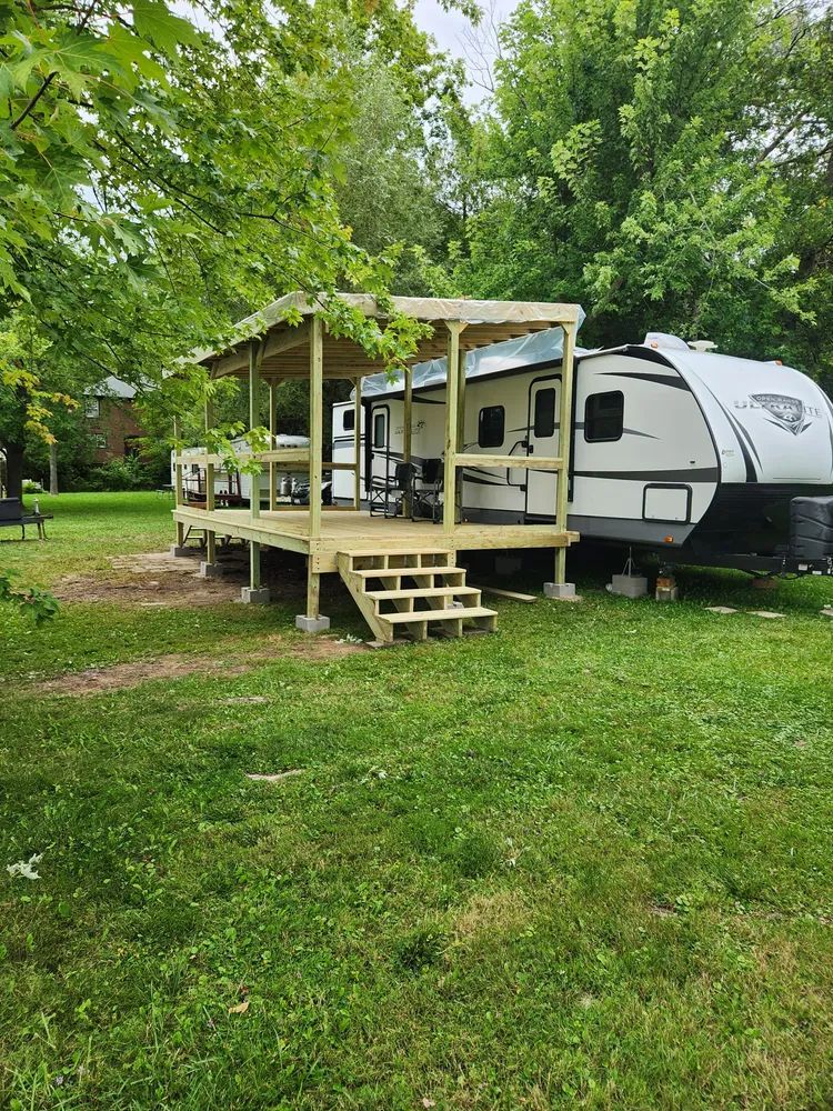 RV with wooden deck and covered area in a grassy yard, surrounded by trees.