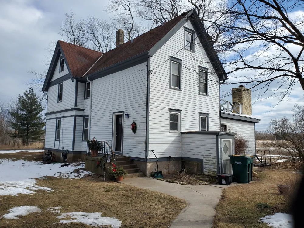 White two-story house with dark roof and trim. Snow-covered ground, leafless trees, overcast sky.