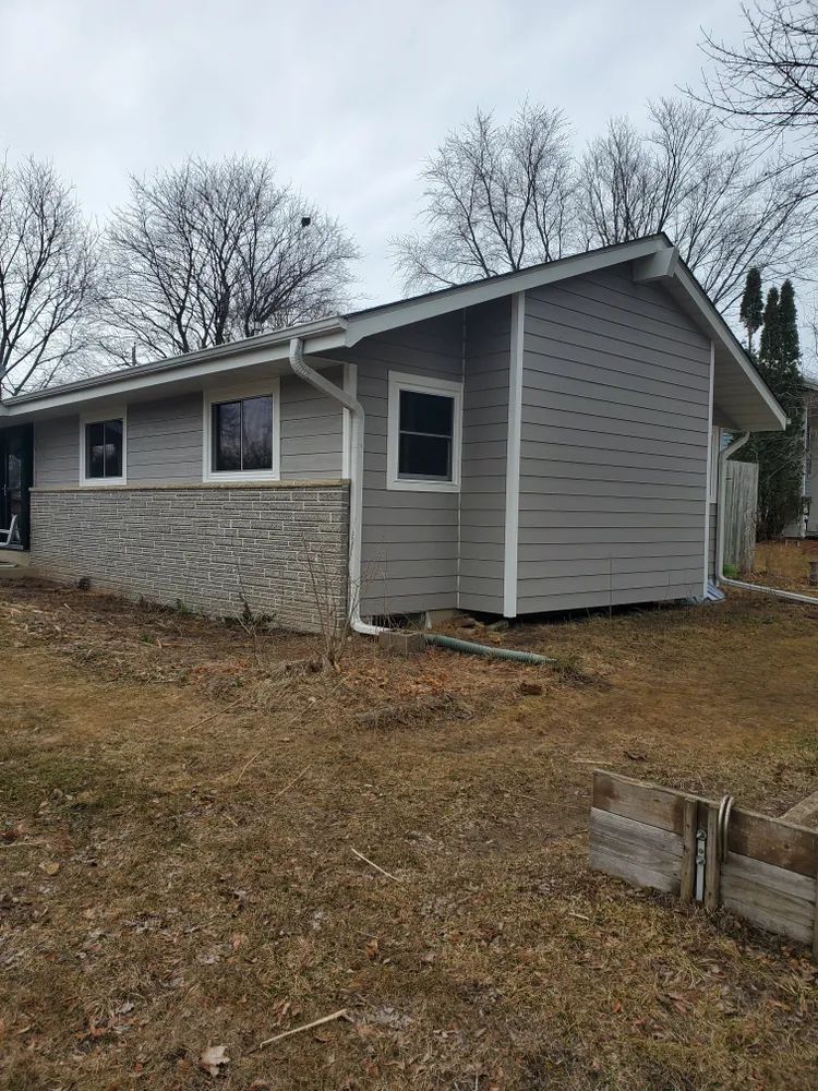 A one-story house with gray siding and stone foundation; bare trees in the background.