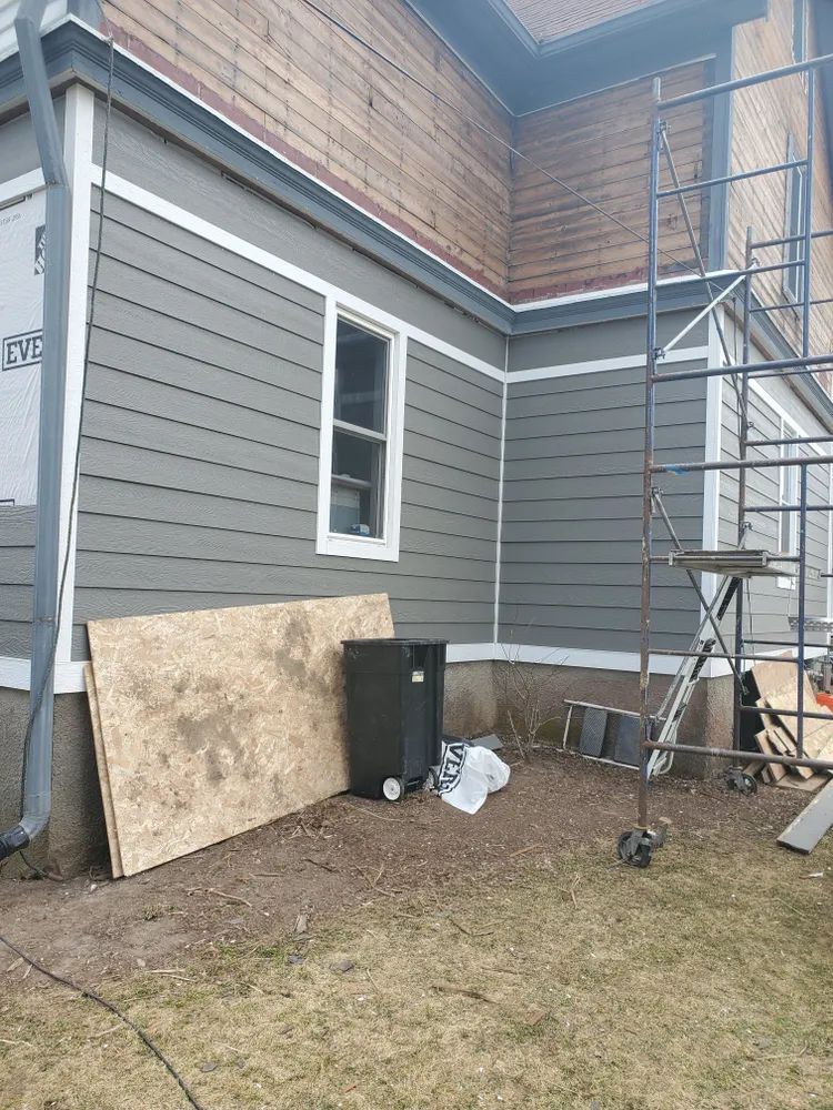 Exterior house with gray siding, white trim, and a window. Scaffolding, debris, and a trash can are present.