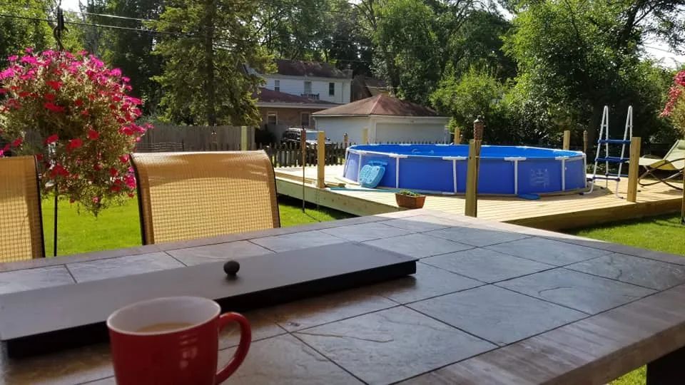 A backyard patio with an above-ground pool and flowers, a table with a coffee cup, and trees in the background.