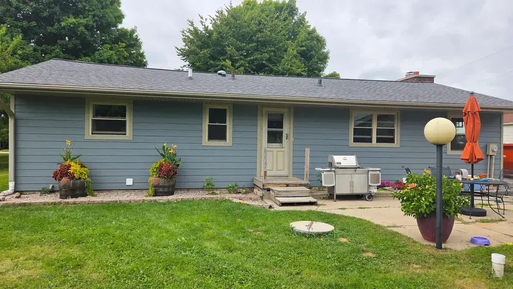 Blue-sided house with a small backyard, green grass, and planters. Gray roof. Overcast day.
