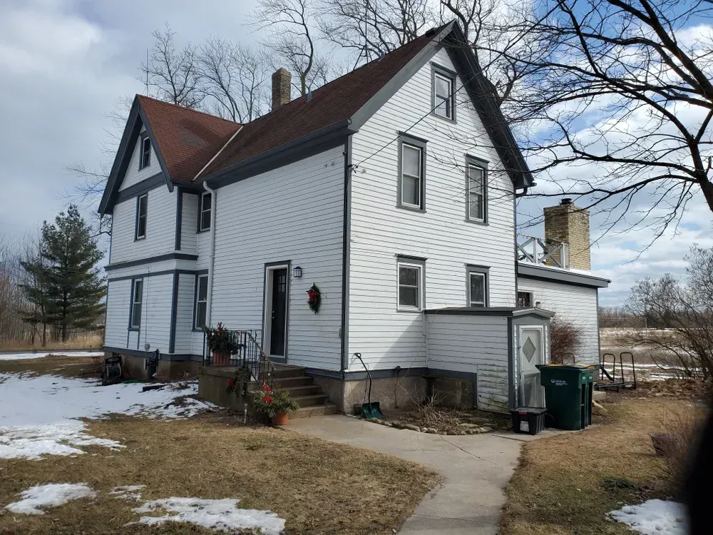 White two-story house with a brown roof and a small concrete walkway. Snow on the ground, trees in background.