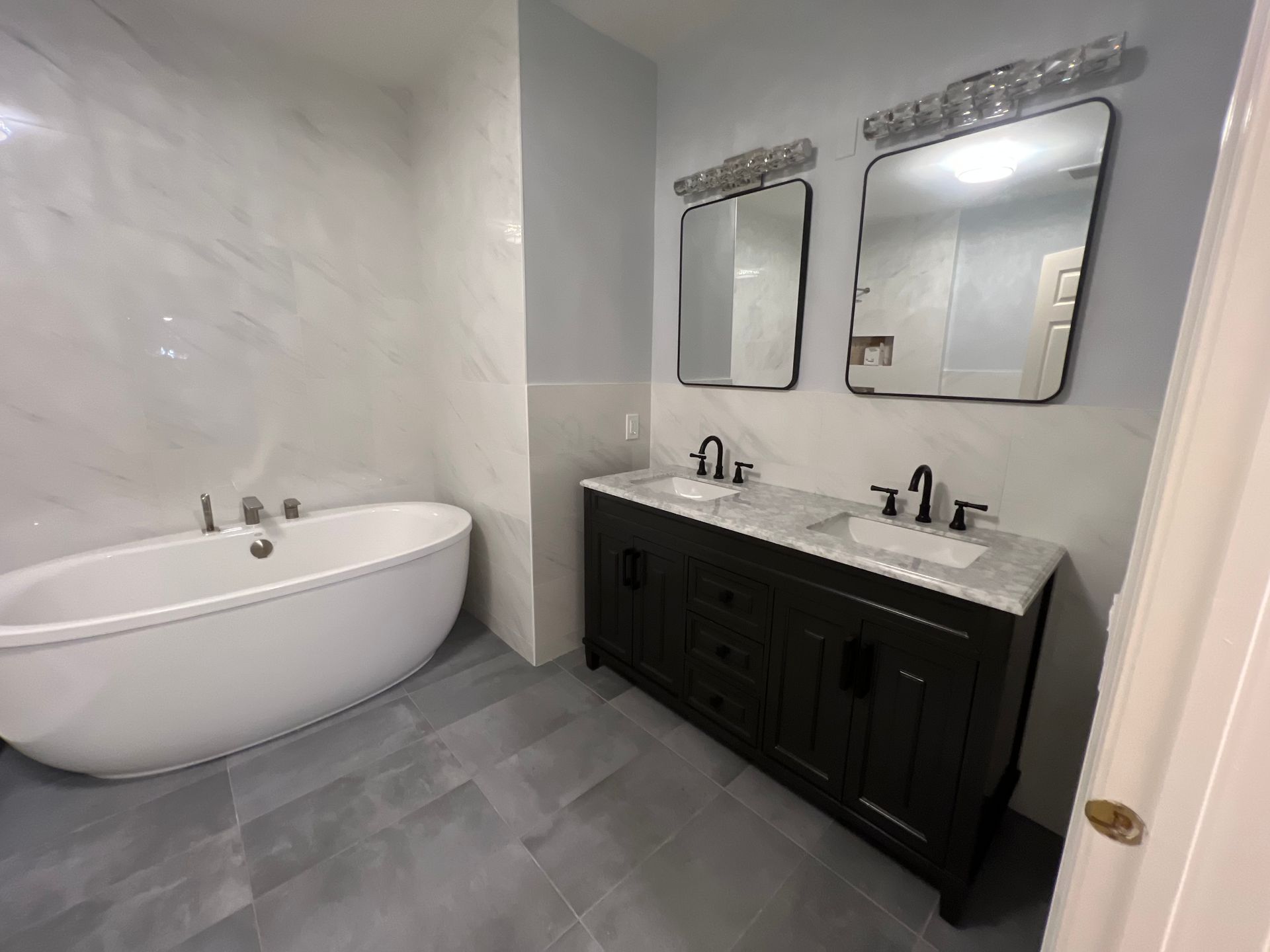 A modern bathroom with a bathtub and double vanity with black fixtures and mirrors. Gray and white color scheme.