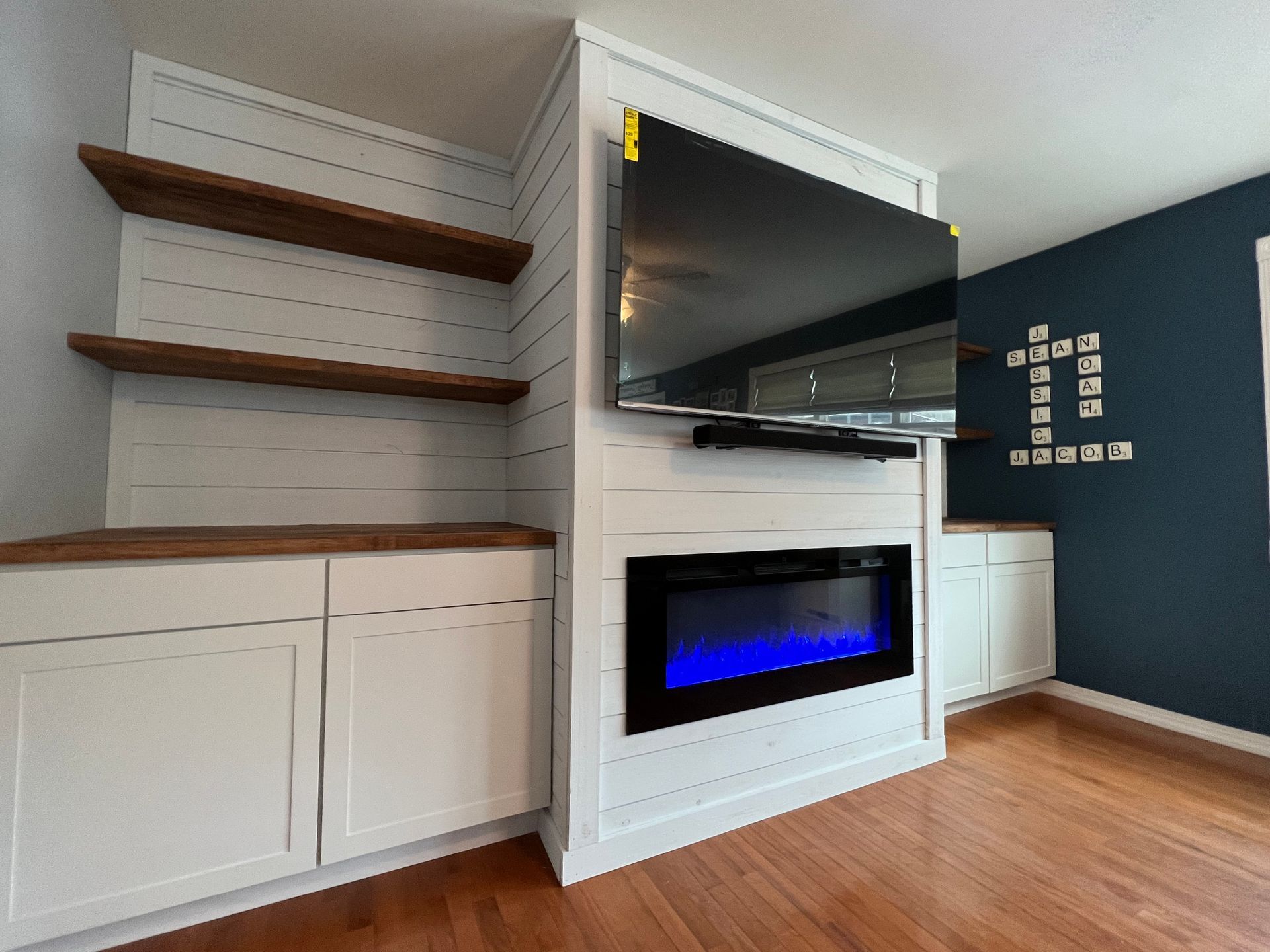 Built-in entertainment center with a fireplace, TV, cabinets, and floating shelves. The wall is white with wood accents.