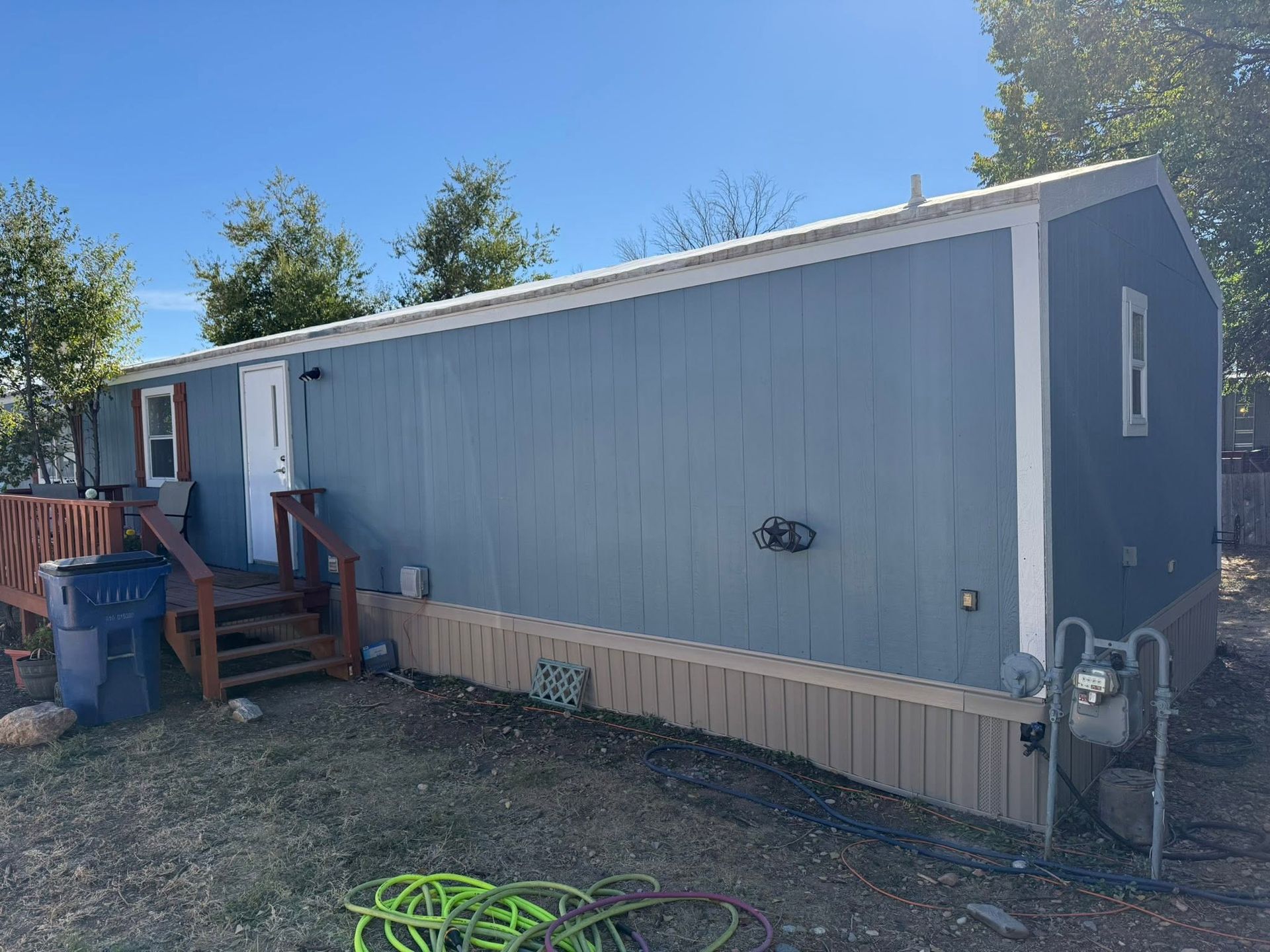 Gray mobile home with a small wooden porch and steps, seen from the side under a clear blue sky.