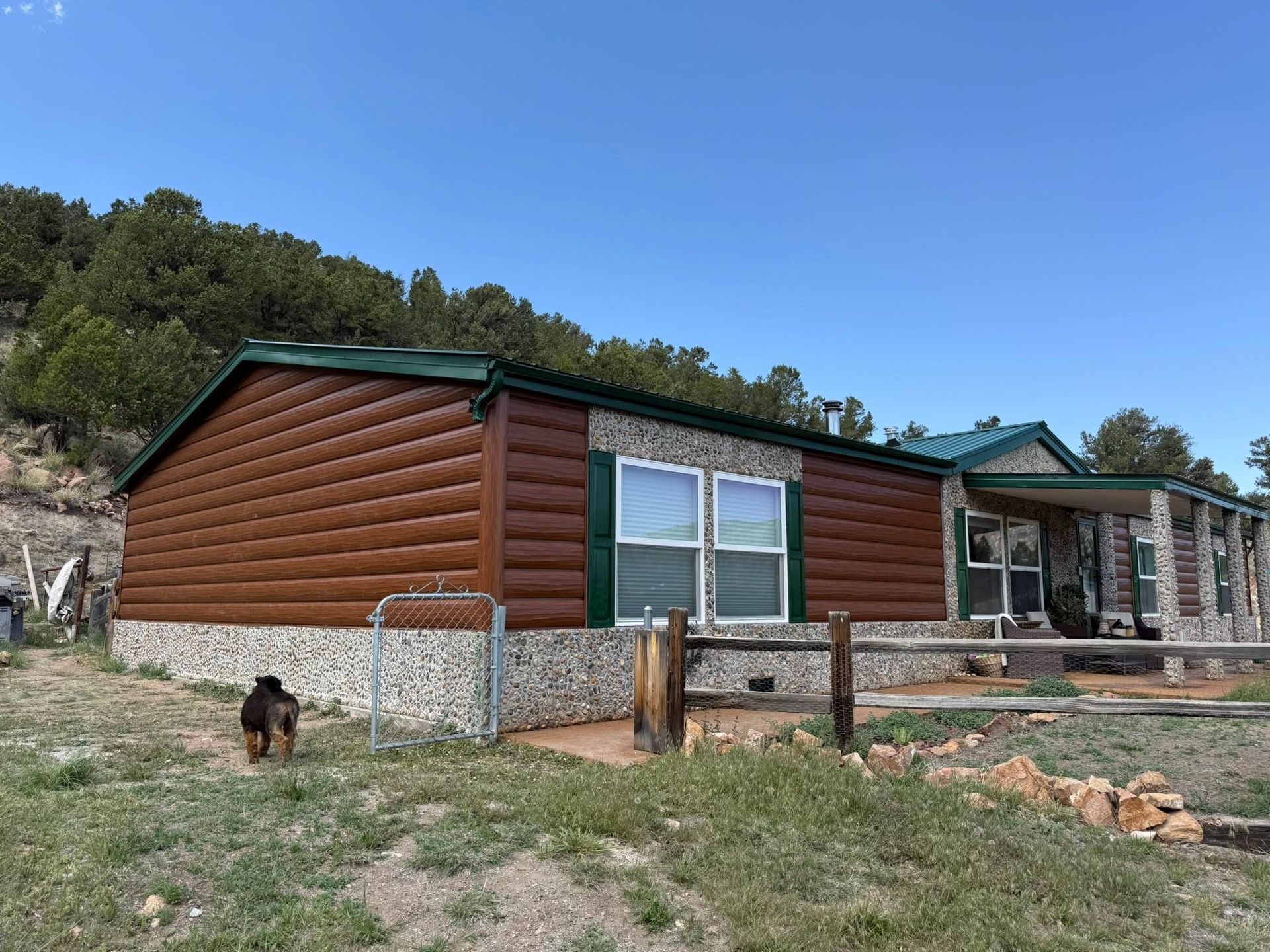 Rustic cabin with green roof and log siding in a rural yard, with a dog and fence in front