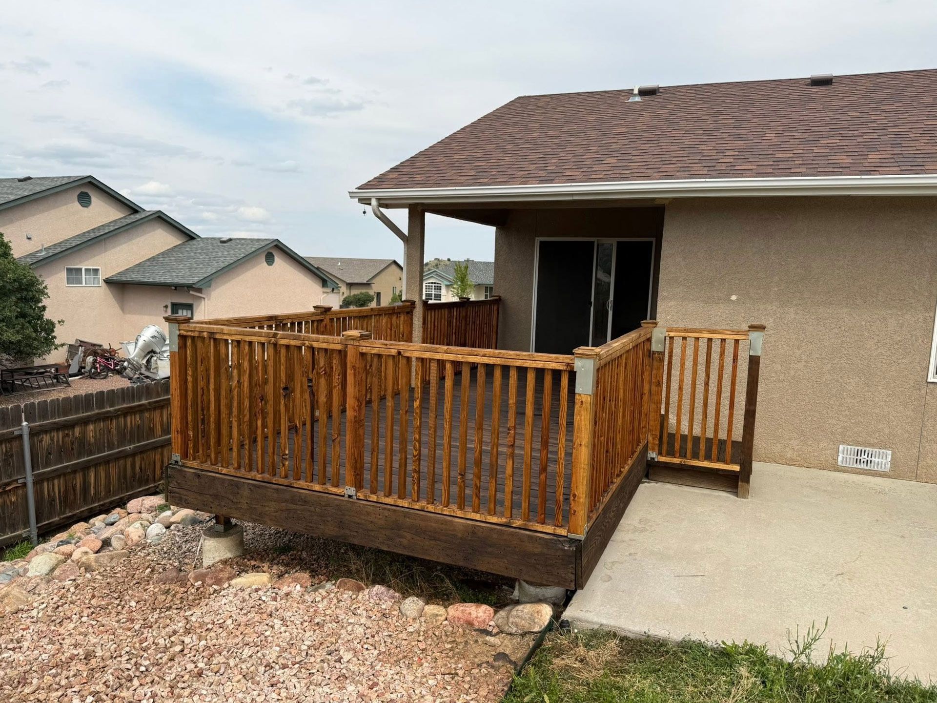 Small wooden deck with railing beside a beige house, next to a concrete patio in a suburban yard