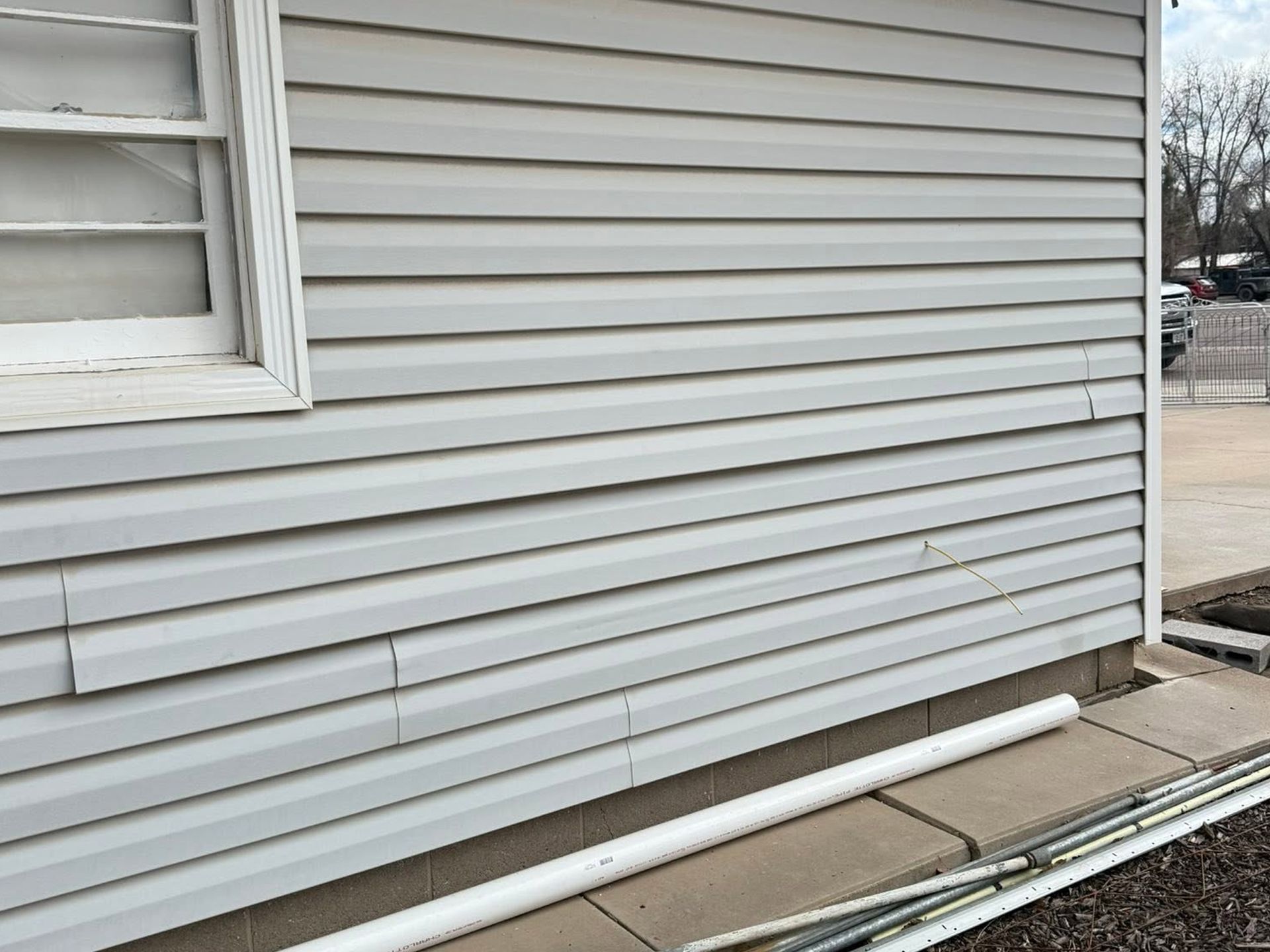 Side of a gray house with vinyl siding and a white pipe lying on the ground near the foundation.