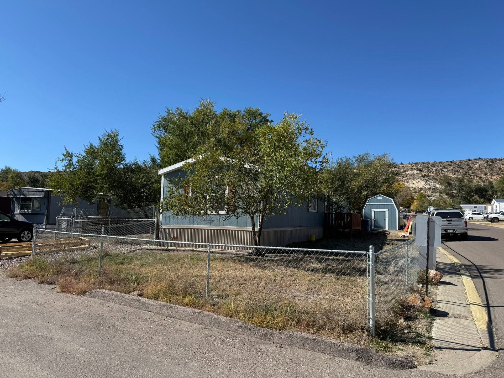 Chain-link fenced lot with trees, small building, and parked vehicles on a sunny roadside.