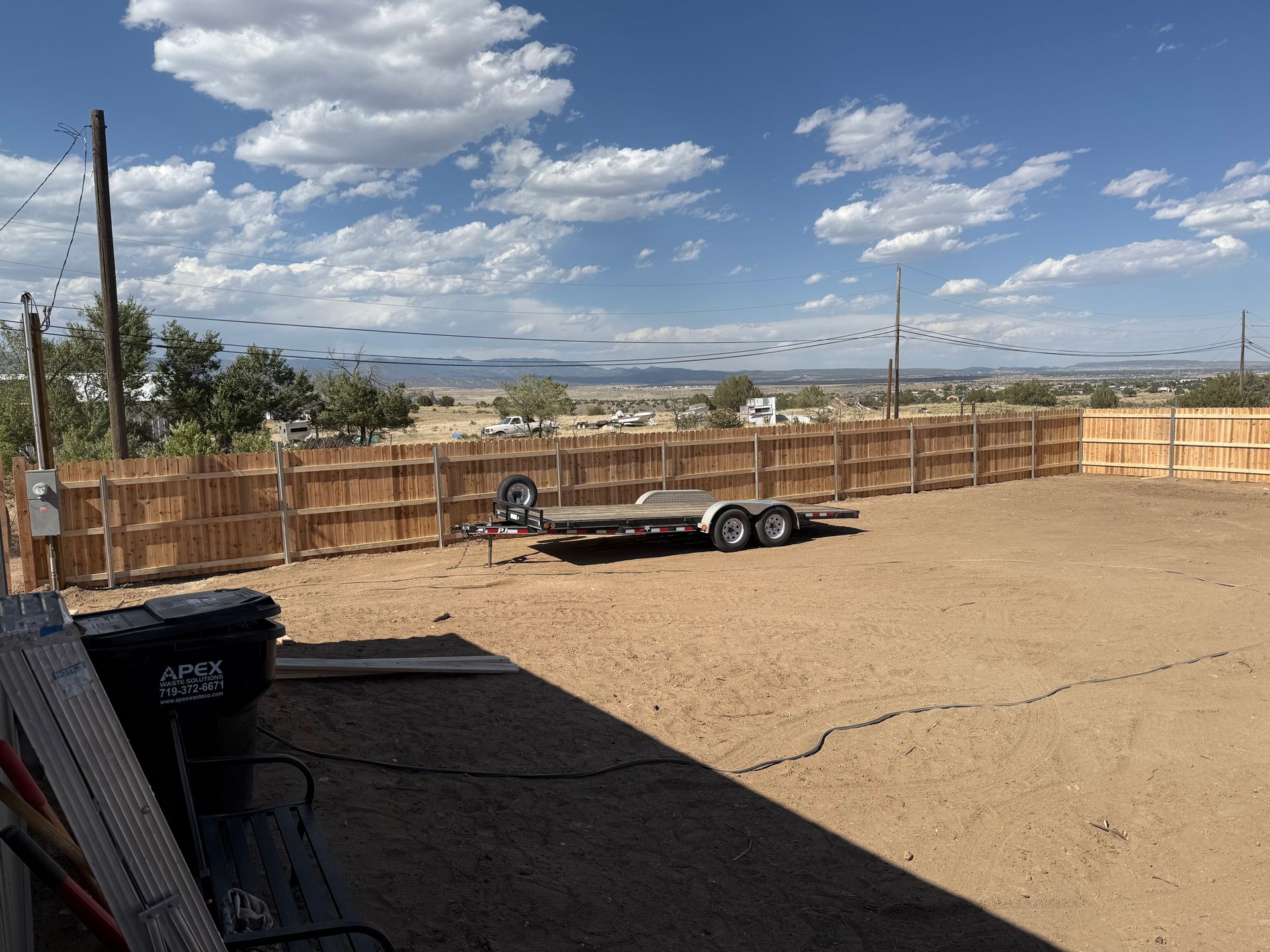 Wide dirt lot with wooden fence, utility trailer, and cloudy blue sky over distant hills