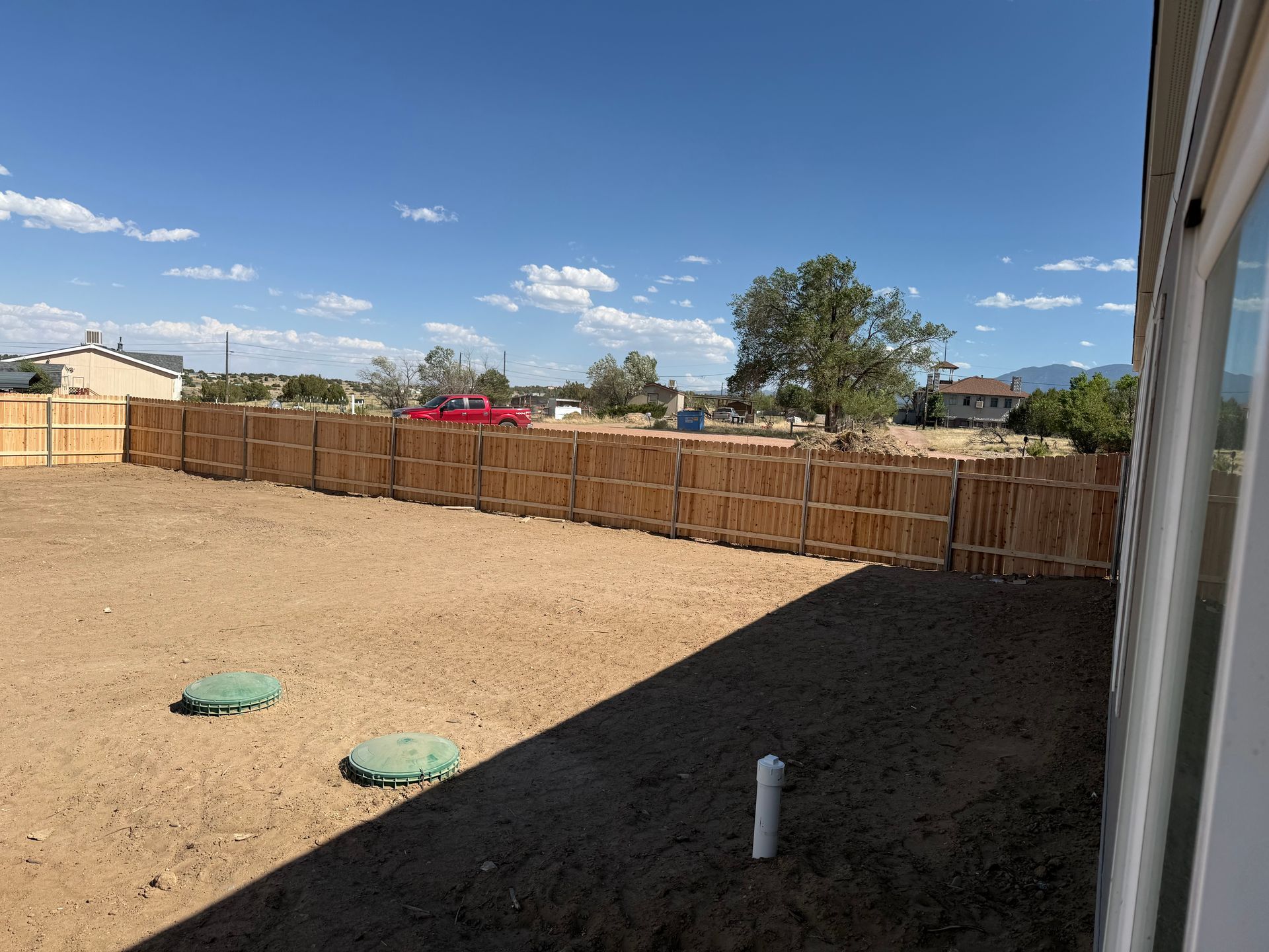 Large dry backyard with a wooden fence, two green covers, and a shaded house wall under a blue sky.