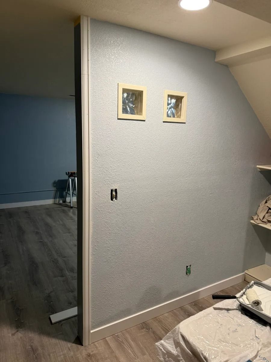 Empty room with gray textured wall, two framed pictures, and partially visible bed or table in foreground