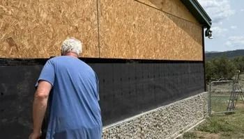 Person in a blue shirt walks beside a building with plywood siding and a black foundation.