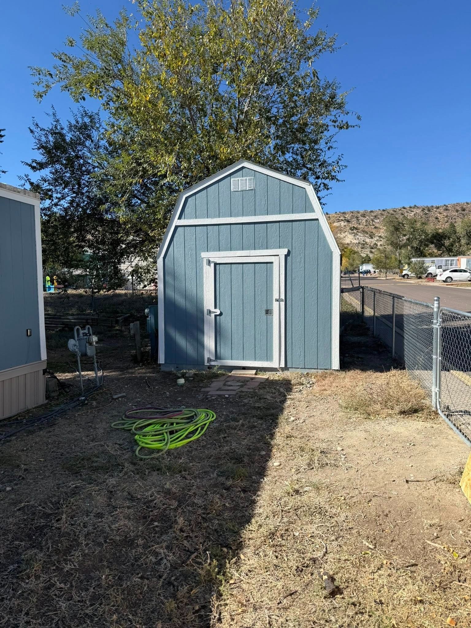 Blue shed with white trim in a backyard, beside a tree and gravel path under a clear sky