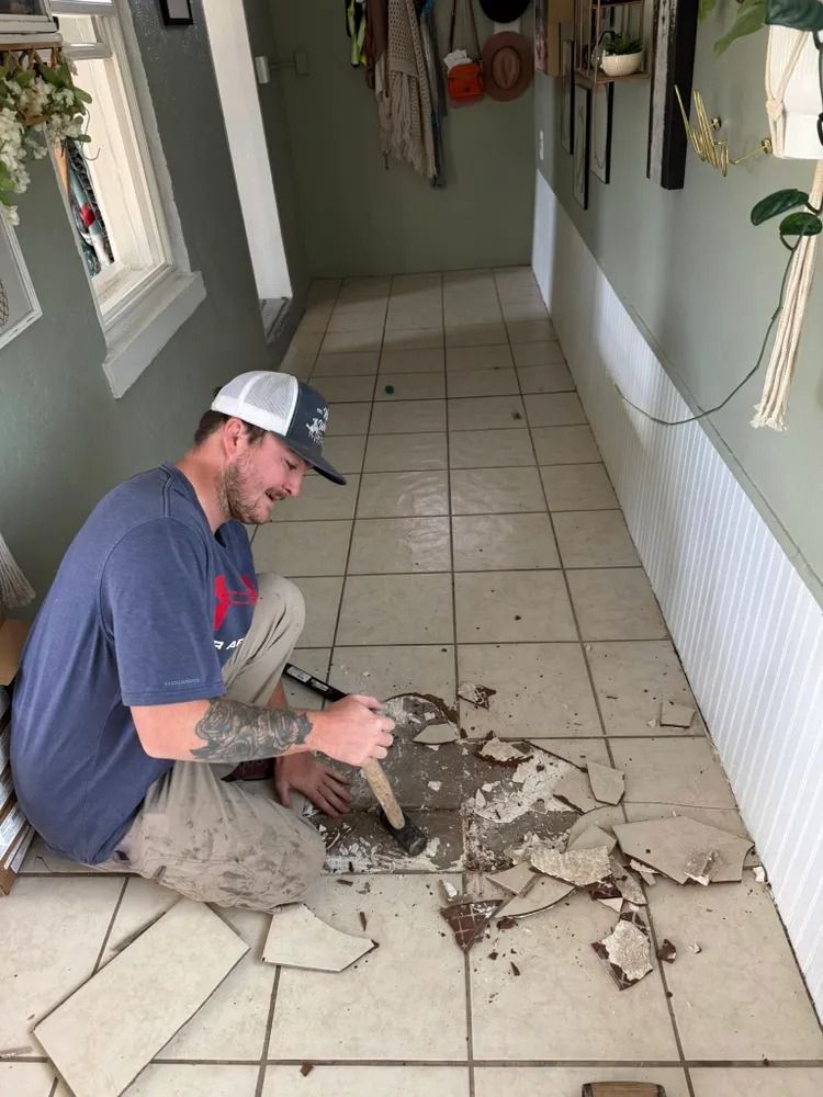 Person kneeling on tiled floor, removing broken tiles in a narrow hallway during renovation