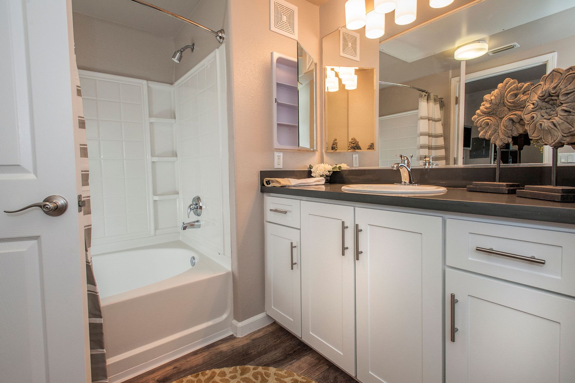 Bathroom in an apartment with white cabinetry, dark countertop, mirror, and a tub/shower.