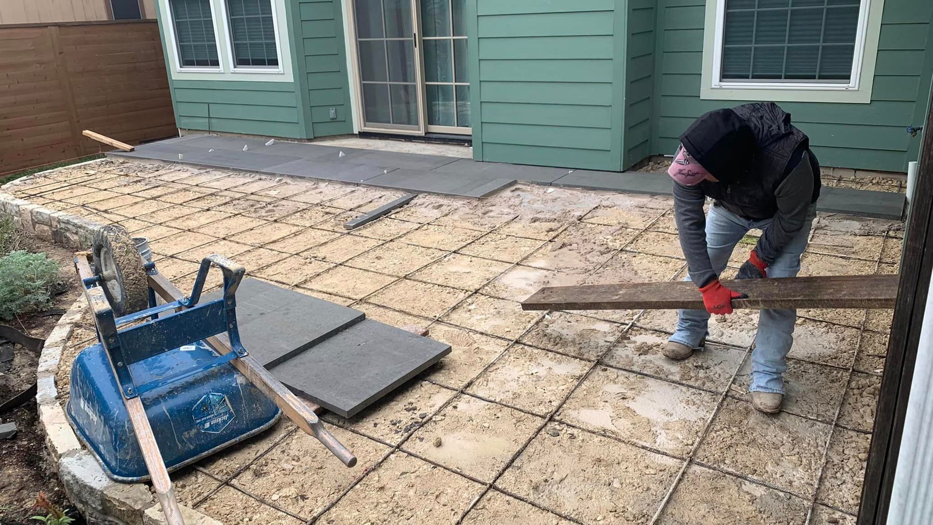 Person in a hooded sweatshirt working on a patio, holding a wooden plank; blue wheelbarrow nearby.