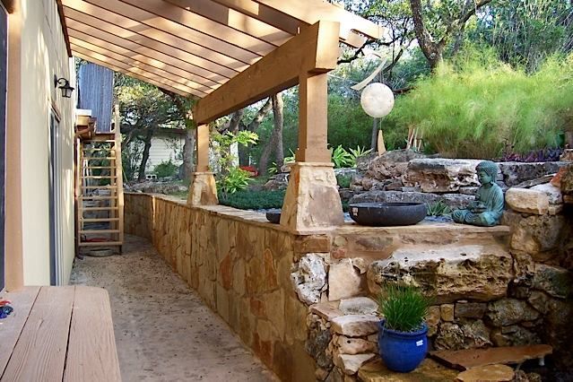 Stone-walled patio with pergola, steps, Buddha statue, and greenery. Brown and beige tones, natural light.