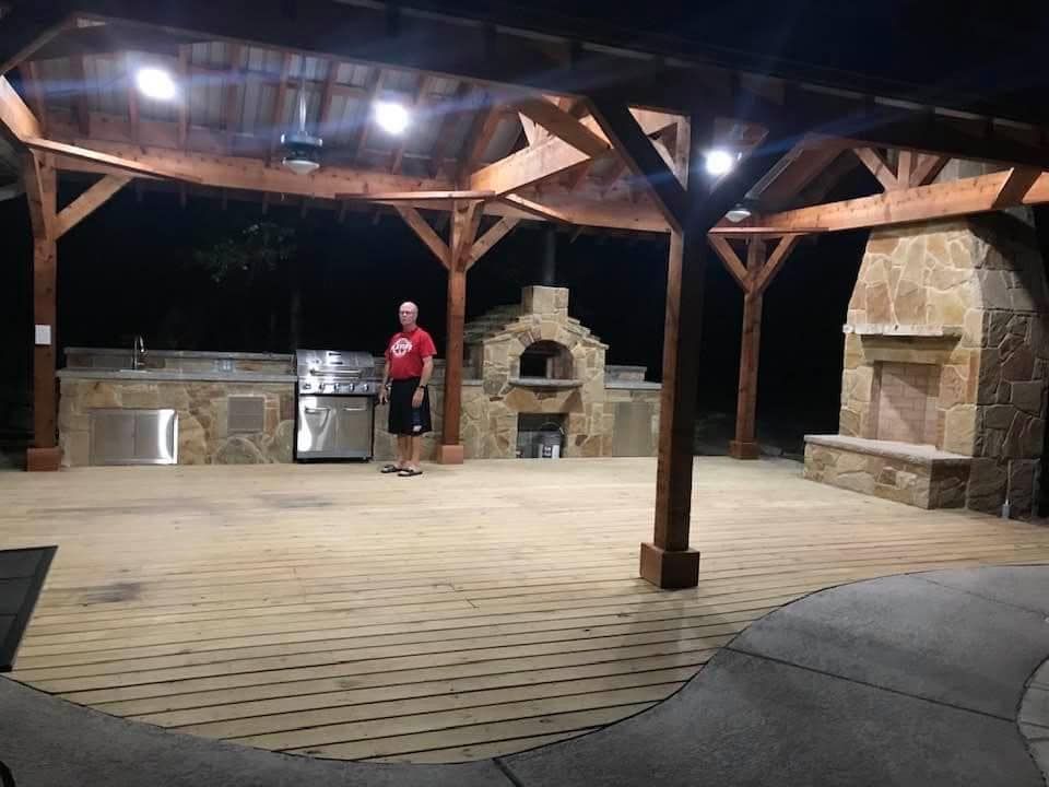 Man standing in outdoor kitchen under a wooden pavilion with fireplace, pizza oven, and built-in grill.