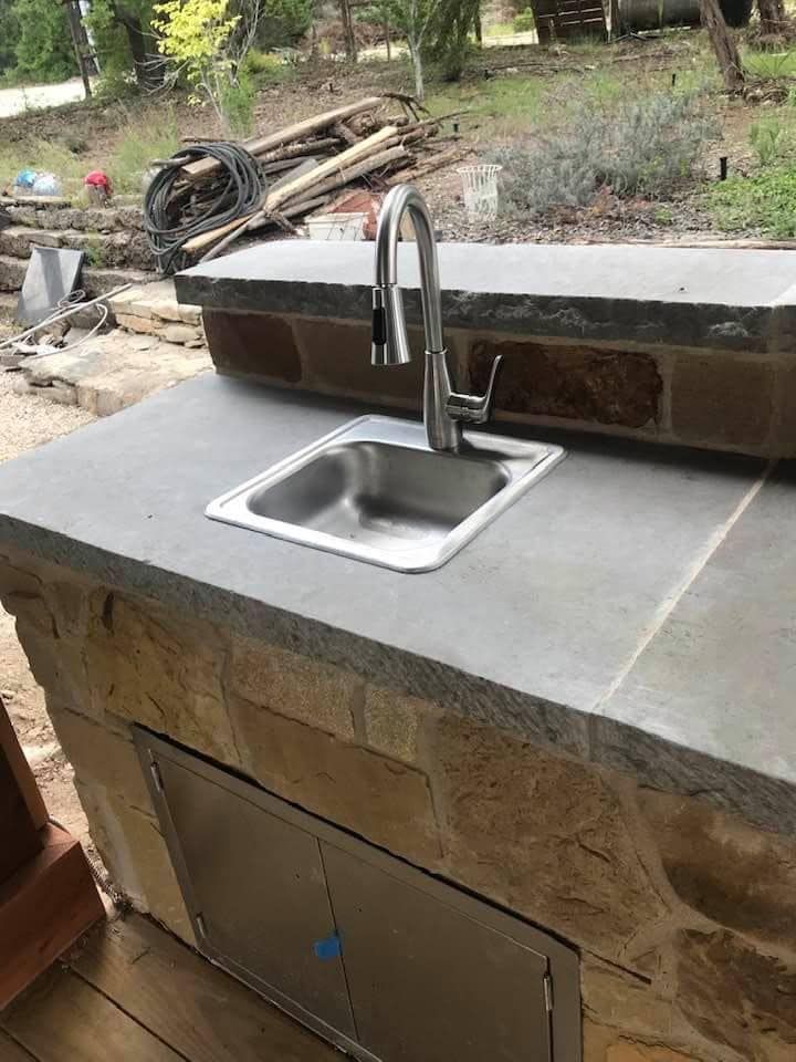 Outdoor kitchen with a stainless steel sink, faucet, stone countertop, and cabinet door.