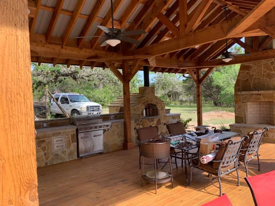 Outdoor kitchen under a wooden canopy with a grill, pizza oven, and fireplace, with a truck parked in the background.