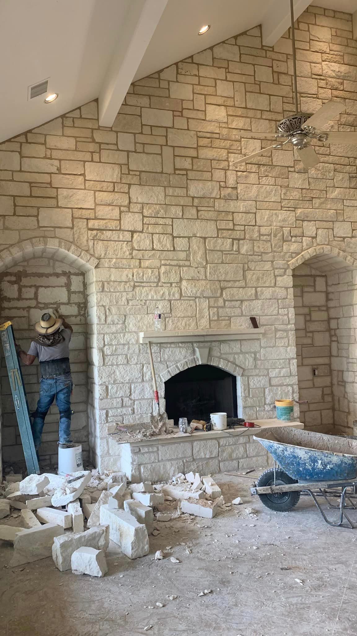 Person working on a stone fireplace with broken stone on the floor. Light stone walls, arched openings.