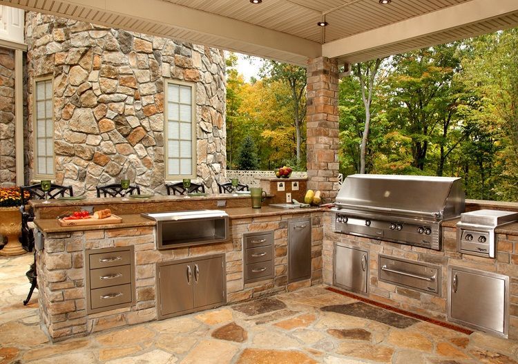 Outdoor stone kitchen with stainless steel appliances, beneath a covered patio with a natural stone floor.