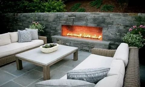 Outdoor seating area with lit fireplace, gray stone wall, white couches, and a coffee table.