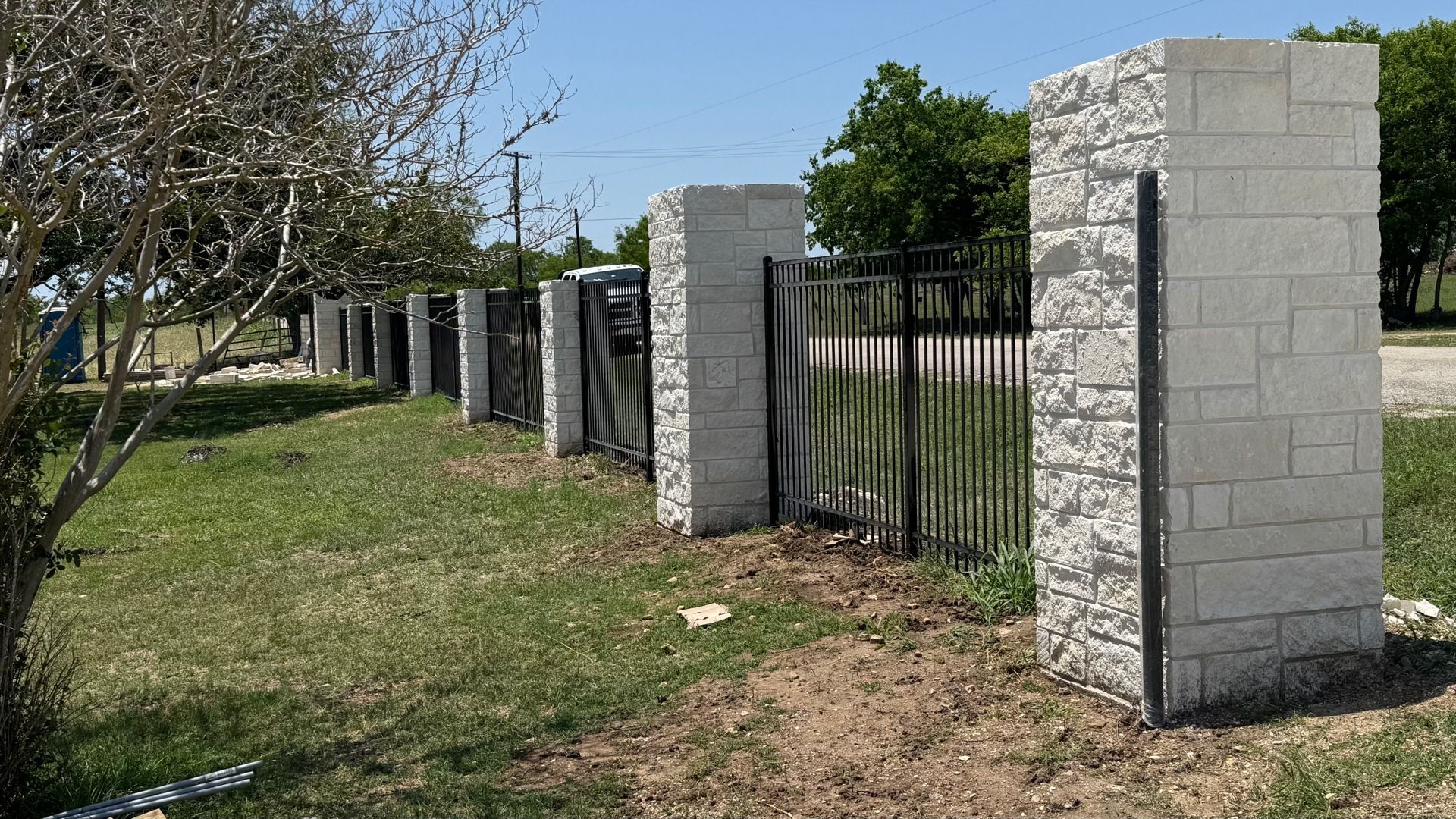 Stone pillars with a black metal fence in a grassy area on a sunny day.