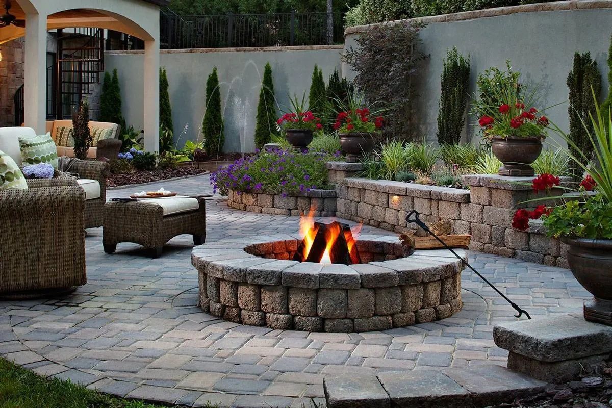 Stone fire pit surrounded by a patio with seating, garden beds, and a wall.