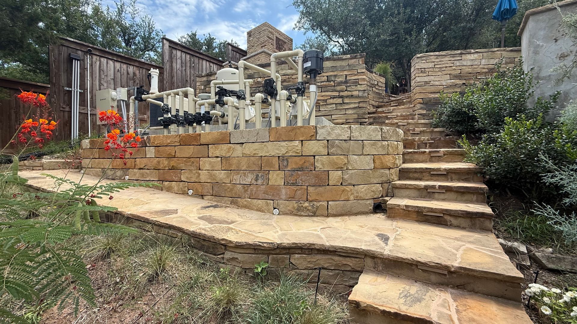Stone retaining wall and steps in a backyard with landscaping and pool equipment visible.