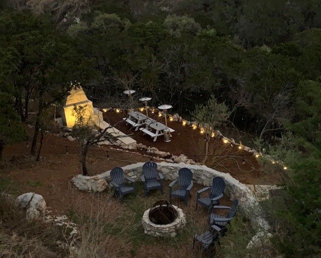 Outdoor dining and seating area with a fire pit, picnic tables, and fairy lights in a wooded setting.
