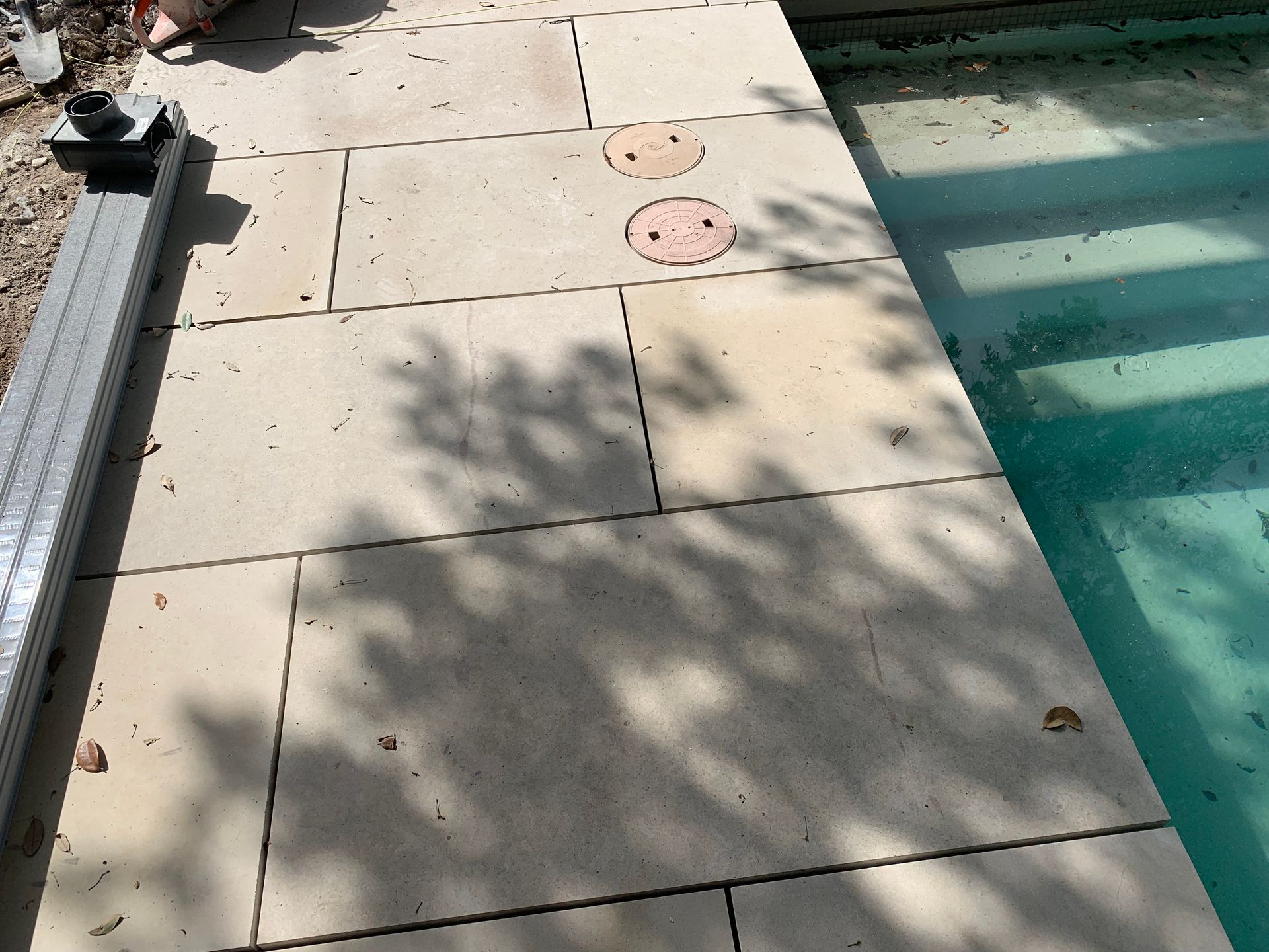Stone patio next to a pool with two circular access covers. Sunlight casts shadows.