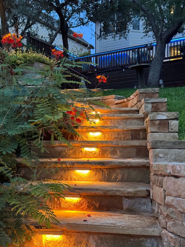 Stone steps with warm lighting leading upward, flowers on either side. Backyard setting at dusk.