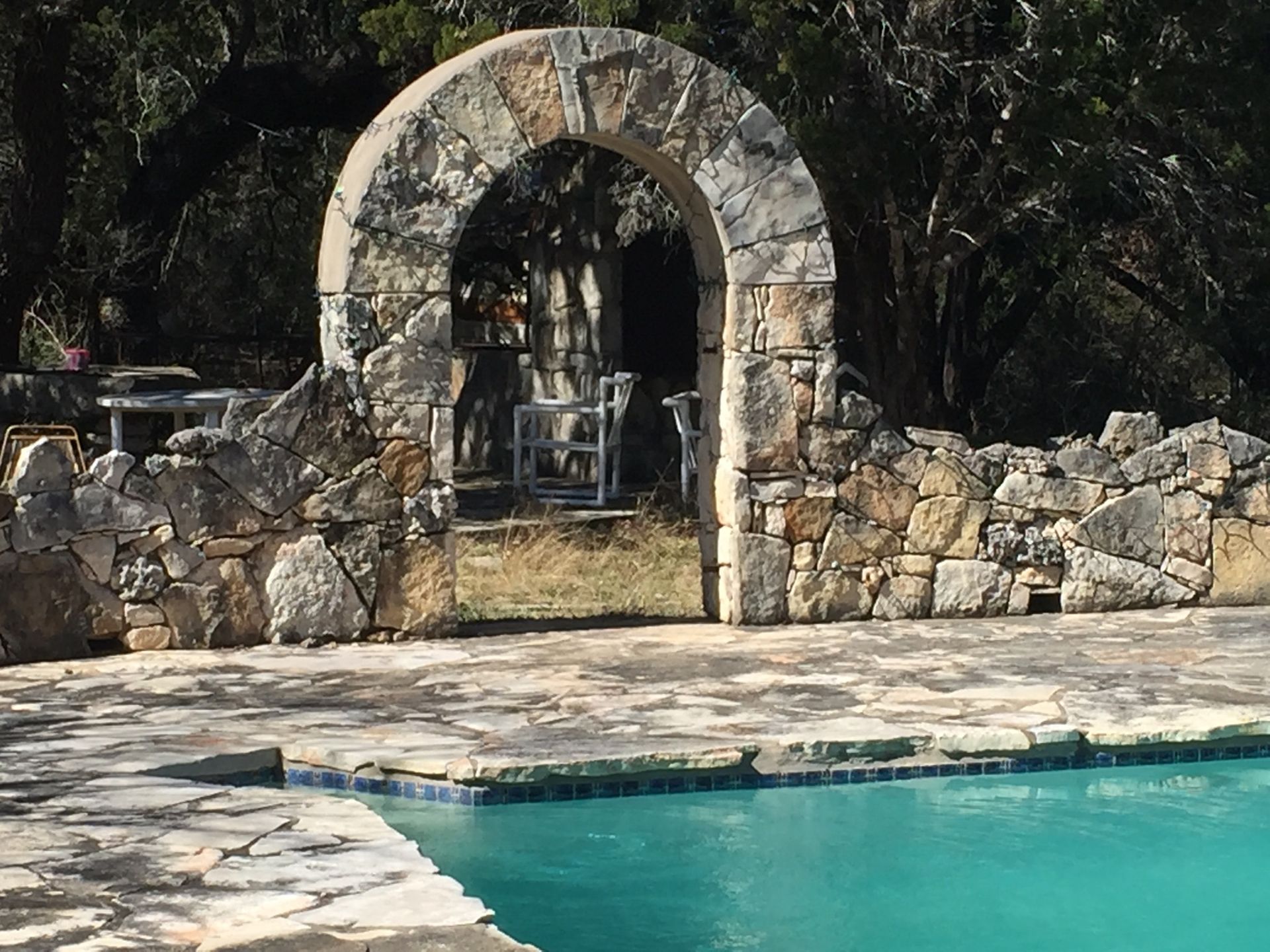 Stone archway frames a pool with teal water. A rock wall and table are visible.
